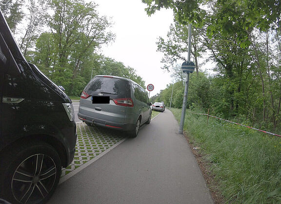 Foto des engen Geh- und Radwegs am Adenauerring. Ein Pkw parkt mit zwei Rädern auf dem Radweg, am rechten Rand steht eine Laterne auf dem Weg.