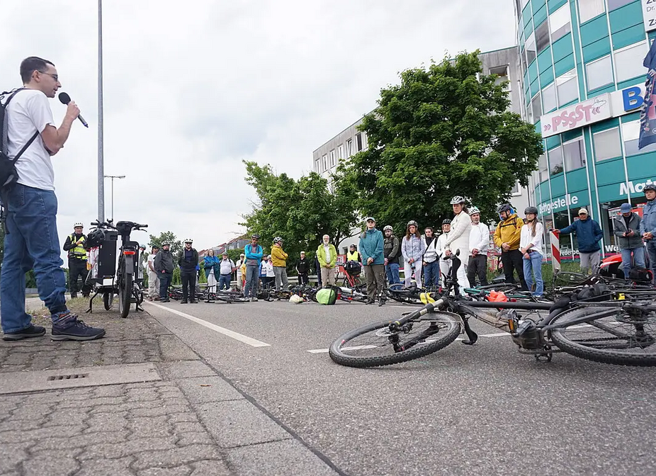 Ride of Silence am 21. Mai 2025 in Karlsruhe, Zwischenkundgebung an der Kreuzung Linkenheimer Landstraße/Moldaustraße Zwischenkundgebung an der Kreuzung Linkenheimer Landstraße/Moldaustraße. Zahlreiche Fahrräder liegen auf der Fahrbahn, Menschen stehen im Kreis darum herum. Am linken Bildrand spricht ein Redner.