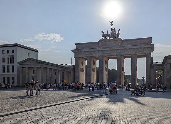 Das Brandenburger Tor in Berlin bei sonnigem Wetter. Im Vordergrund ist ein großer, gepflasterter Platz zu sehen, auf dem sich Menschen aufhalten. Einige fahren mit Fahrrädern, andere spazieren oder stehen in Gruppen zusammen. Die Quadriga auf dem Tor ist von der Sonne im Hintergrund leicht überstrahlt.