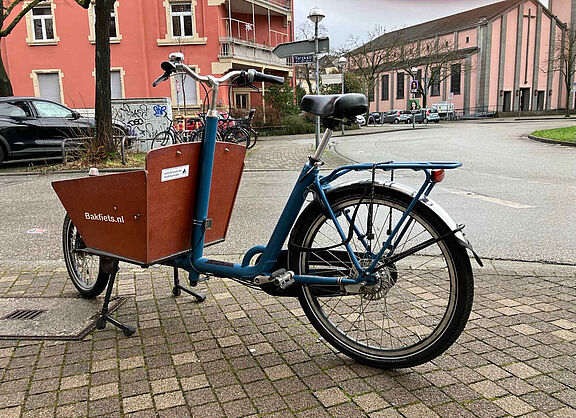 Foto eines abgestellten Lastenrads Typ Bakfiets Classic Short ohne Motor. Auf der hölzernen Kiste ist ein Aufkleber angebracht, der auf die Förderung der Stadt Karlsruhe hinweist.