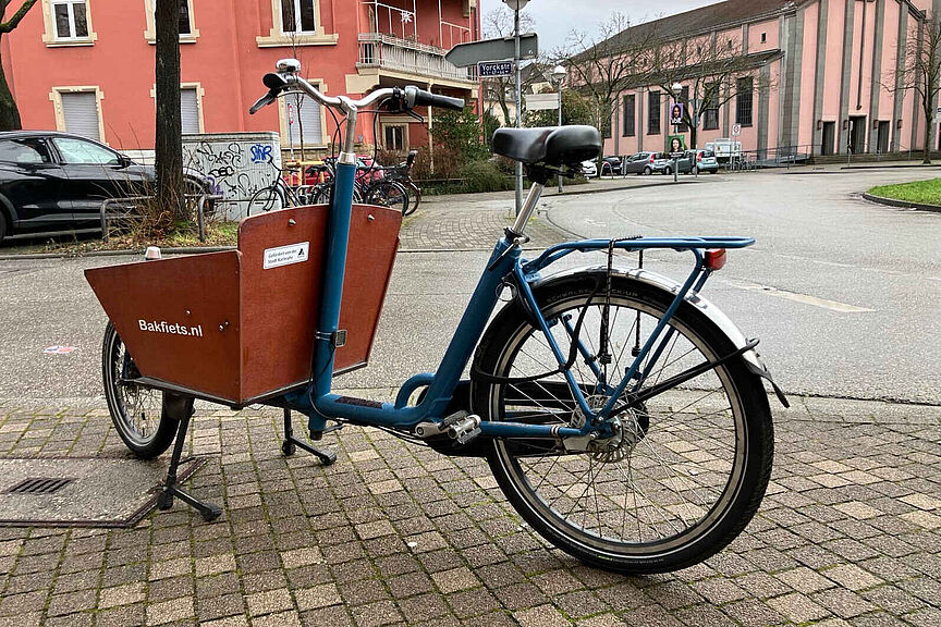 von der Stadt Karlsruhe gefördertes Lastenrad Foto eines abgestellten Lastenrads Typ Bakfiets Classic Short ohne Motor. Auf der hölzernen Kiste ist ein Aufkleber angebracht, der auf die Förderung der Stadt Karlsruhe hinweist.