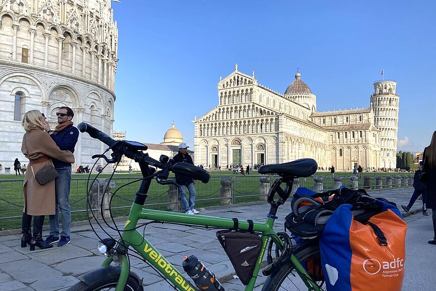Im Vordergrund steht ein grünes Faltrad mit Gepäcktaschen, darunter eine orange blaue Tasche mit ADFC Logo. Im Hintergrund sind der Domplatz in Pisa mit dem Dom, dem schiefen Turm und ein Teil des Baptisteriums zu sehen, außerdem mehrere Besucher:innen auf der Wiese und auf dem Weg bei blauem Himmel.