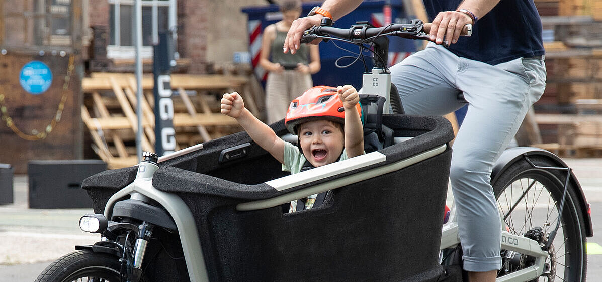 Ein Kind in einem Lastenrad hebt jubelnd die Arme, das Lastenrad fährt eine erwachsene Person in Jeans. für den Kindertransport ist der Traum vieler junger Familien.
