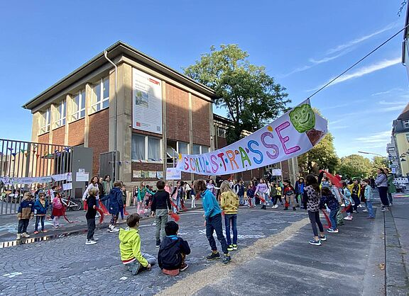 Schulstraße Kinder verschiedenen Alters spielen und bewegen sich auf einer gepflasterten Straße vor einem zweistöckigen Backsteingebäude. Über der Straße hängt ein buntes Banner mit der Aufschrift „Schulstraße“ und einem grünen Baum-Symbol. Im Hintergrund sind Erwachsene zu sehen, die die Aktion begleiten.