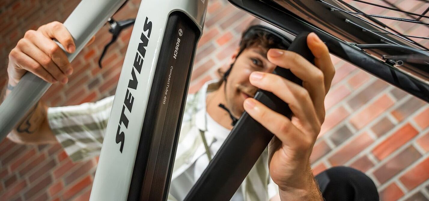 Akku Ein Mann öffnet die Akku-Abdeckung an einem grauen Elektrorad, von schräg unten aufgenommen.